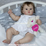 Boneca bebê reborn realista Catharina deitada em berço branco com travesseiro cinza estampado de coroas, vestida de branco e segurando ursinho de pelúcia.