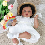 Boneca Reborn Cecília de pele negra deitada sobre tapete felpudo bege, vestindo vestido branco de renda, segurando borboleta decorativa e usando chapéu de palha.