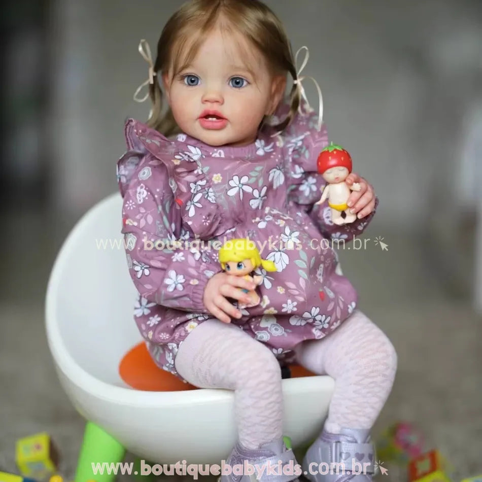 Boneca Bebê Reborn Julia sentada em cadeirinha branca com brinquedos nas mãos, vestindo vestido floral lilás e meia calça.