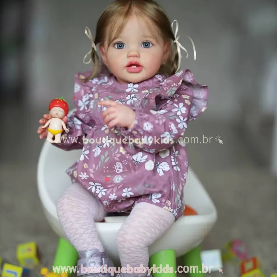 Boneca Bebê Reborn Julia com vestido floral roxo, segurando dois brinquedos e sentada em cadeirinha infantil.