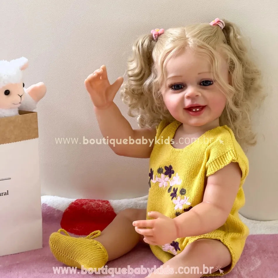 Boneca reborn Lara sorrindo e acenando, sentada sobre toalha rosa ao lado de uma caixa com pelúcia de ovelha.