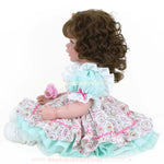 Boneca Bebê Reborn Maria de perfil sentada, segurando chupeta, com vestido estampado e cabelo cacheado.