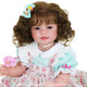 Boneca Bebê Reborn Maria sorrindo, com cabelo solto cacheado e laço azul com detalhes coloridos.