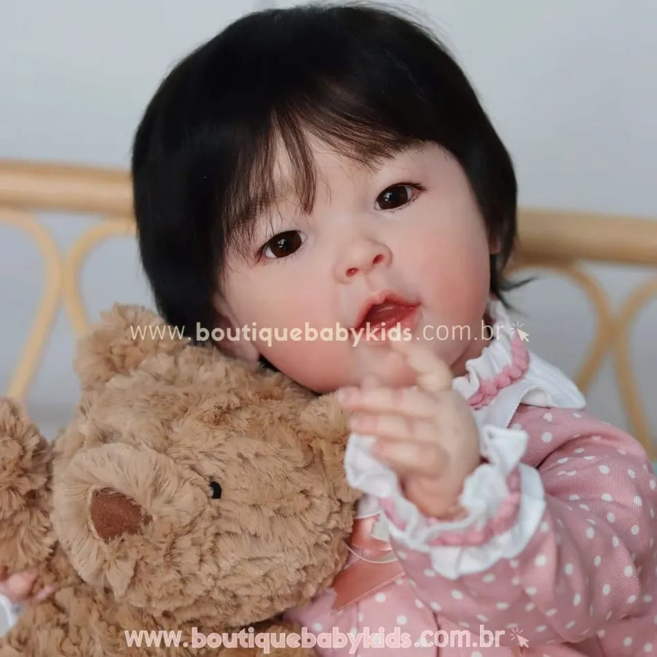 Close da boneca bebê reborn asiática com touca rosa de poá, abraçando um ursinho fofo.