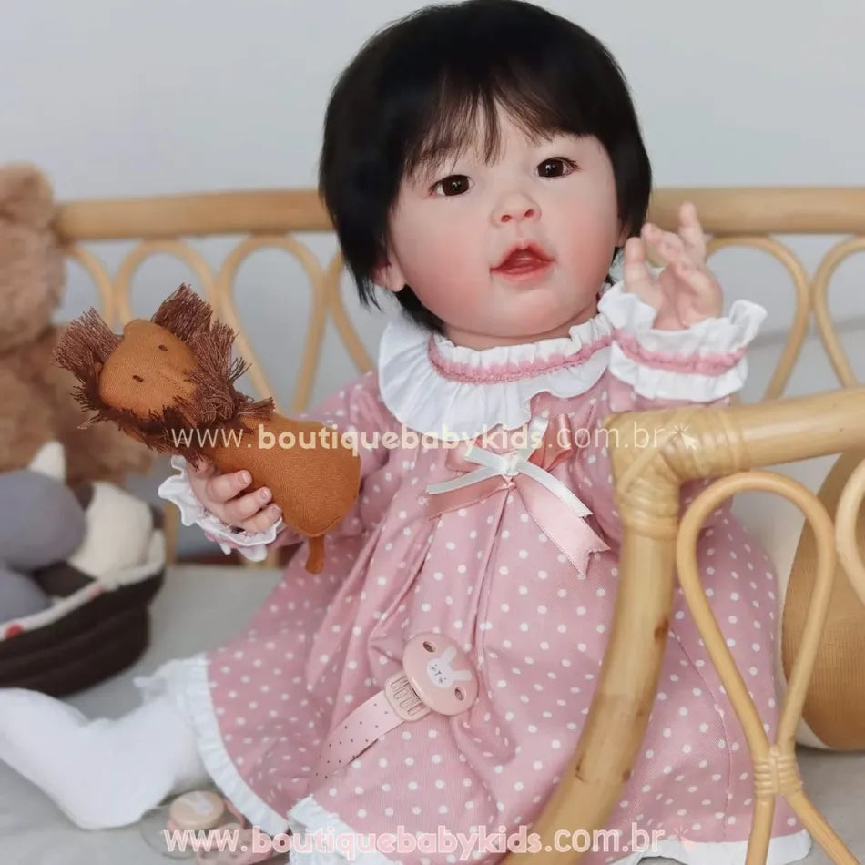 Close da boneca bebê reborn realista asiática com touca rosa e expressão delicada, segurando um ursinho de pelúcia.