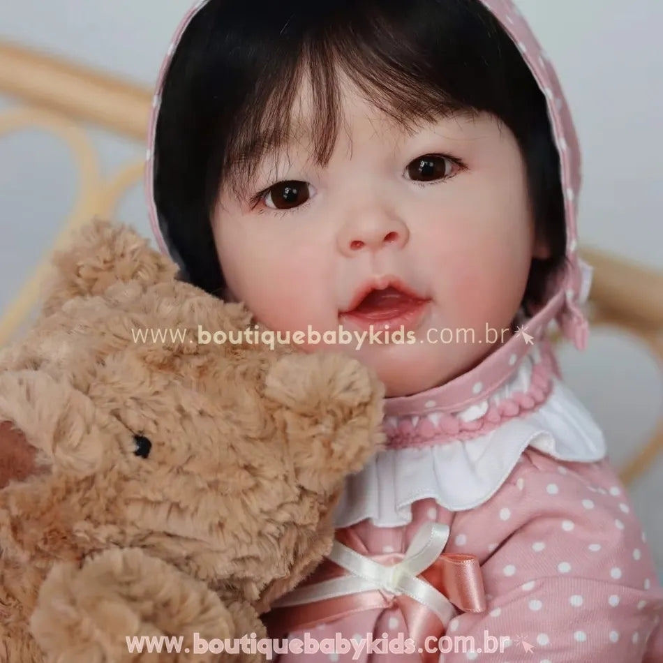 Boneca bebê reborn realista asiática com touquinha rosa de poá, chupeta magnética e urso de pelúcia no colo.