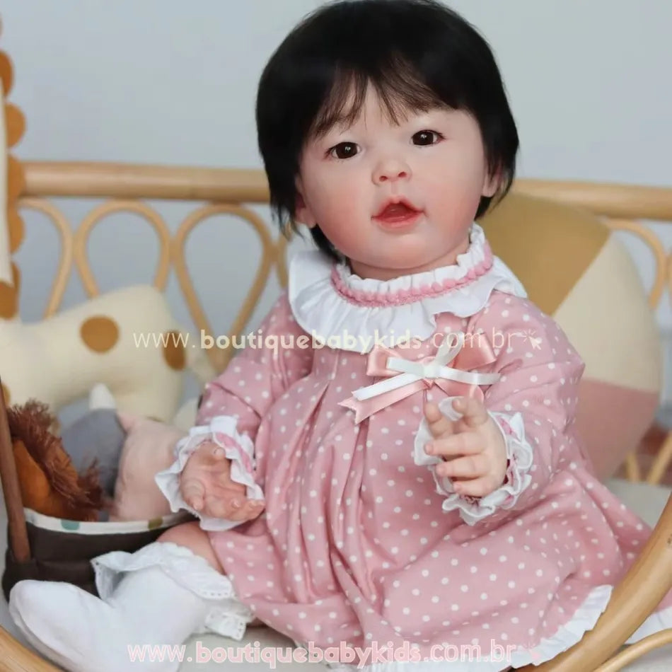 Boneca bebê reborn realista asiática com cabelo preto liso, vestindo vestido rosa com poás brancos e gola branca, sentada em uma cadeira de vime.