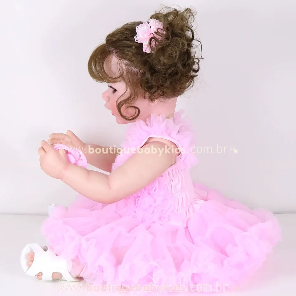 Boneca Reborn Rebecca de perfil, segurando chupeta rosa com as duas mãos, com vestido rosa e cabelo cacheado preso