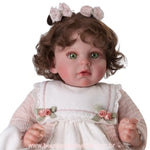 Retrato em close da Boneca Bebê Reborn destacando expressão facial realista, laços no cabelo e detalhes de flores de cetim no bolero de tricô rosa.