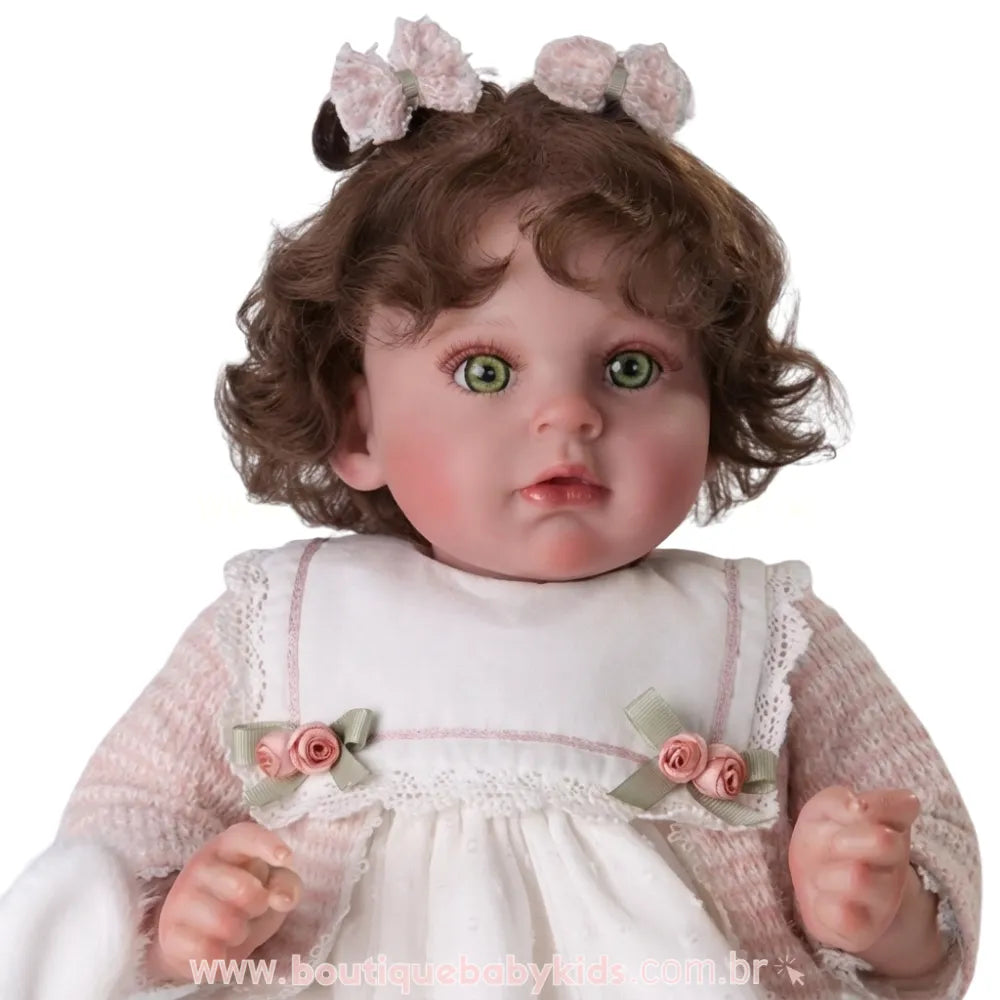Retrato em close da Boneca Bebê Reborn destacando expressão facial realista, laços no cabelo e detalhes de flores de cetim no bolero de tricô rosa.