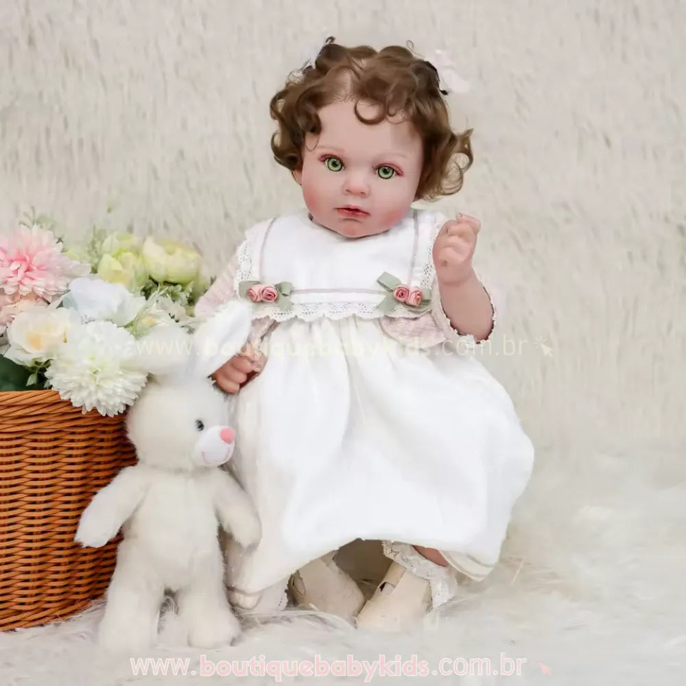 Boneca realista estilo Reborn sentada ao lado de uma cesta de vime com flores artificiais coloridas, vestindo conjunto clássico branco e rosa, com coelho de pelúcia.