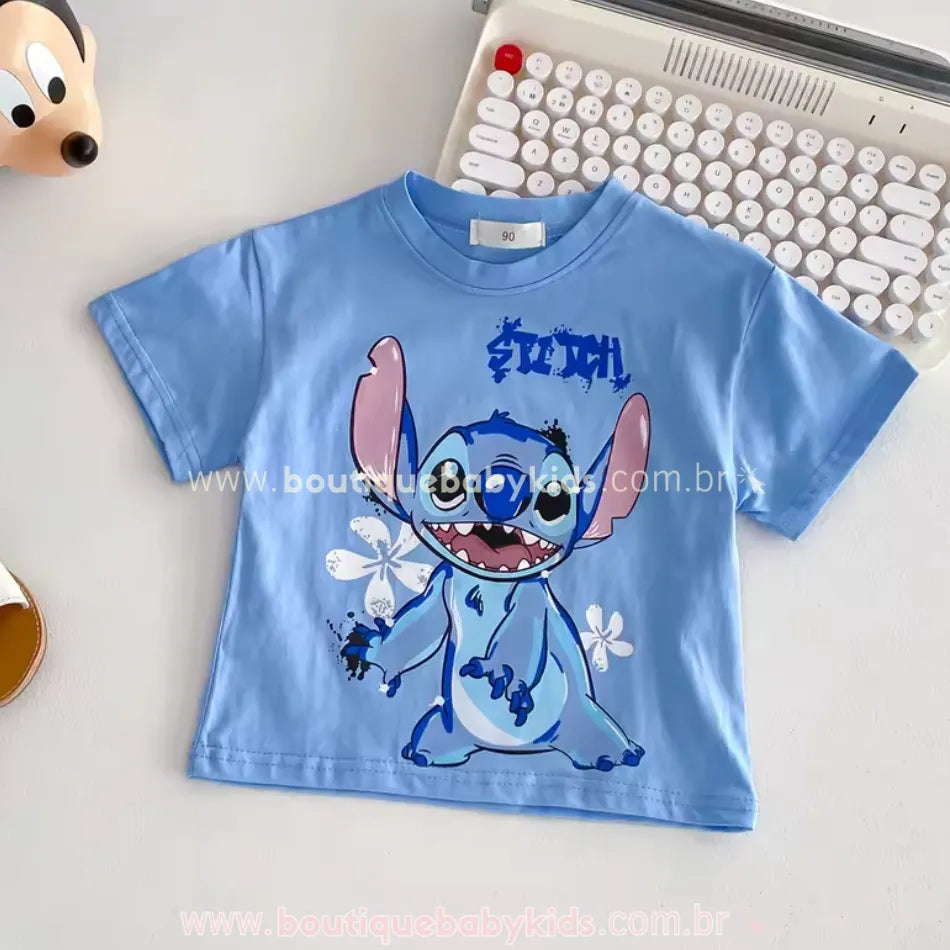 Camiseta infantil azul com estampa do personagem Stitch sorrindo, disposta sobre superfície clara ao lado de uma máquina de escrever branca e brinquedo do mickey.