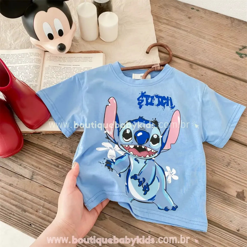 Camiseta azul infantil com estampa do Stitch, sendo segurada por uma mão, com fundo de madeira, livro aberto, bota vermelha e enfeite do Mickey Mouse ao fundo.