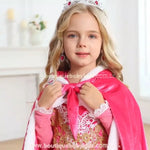 Menina sorridente vestindo a capa rosa, segurando o laço de amarração frontal enquanto posa com um visual digno de realeza, evidenciando os detalhes sofisticados da peça.