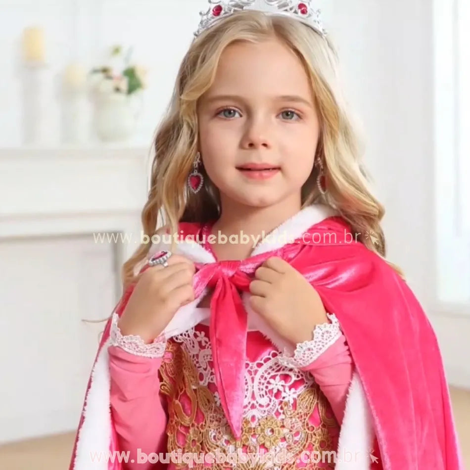 Menina sorridente vestindo a capa rosa, segurando o laço de amarração frontal enquanto posa com um visual digno de realeza, evidenciando os detalhes sofisticados da peça.