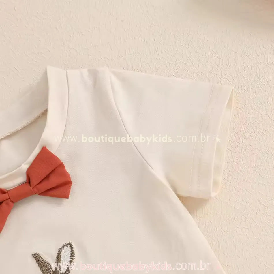 Detalhe da manga curta da camiseta infantil bege com gola arredondada e laço decorativo em tom terroso. Peça feita em algodão, macia e confortável para o bebê.