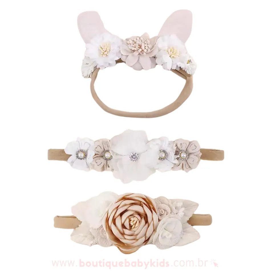 Três faixas para bebê com detalhes florais e pérolas, incluindo uma faixa com orelhinhas de coelho e flores brancas e bege, uma faixa delicada com flores de tecido e pérolas e uma faixa sofisticada com uma flor grande central de cetim bege.