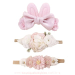 Kit com três faixas florais delicadas, incluindo uma faixa com orelhinhas de coelho em tecido rosa e laço de cetim, uma faixa com flores brancas e rosadas em camadas e uma faixa decorada com flores de cetim e pequenos detalhes brilhantes.