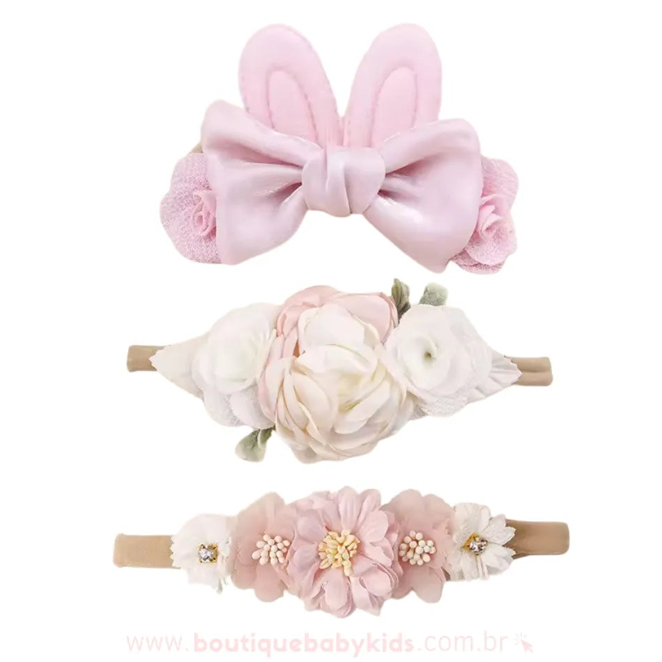 Kit com três faixas florais delicadas, incluindo uma faixa com orelhinhas de coelho em tecido rosa e laço de cetim, uma faixa com flores brancas e rosadas em camadas e uma faixa decorada com flores de cetim e pequenos detalhes brilhantes.
