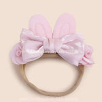 Faixa infantil rosa com orelhinhas de coelho acolchoadas e laço de cetim no centro, decorada com pequenas flores laterais.