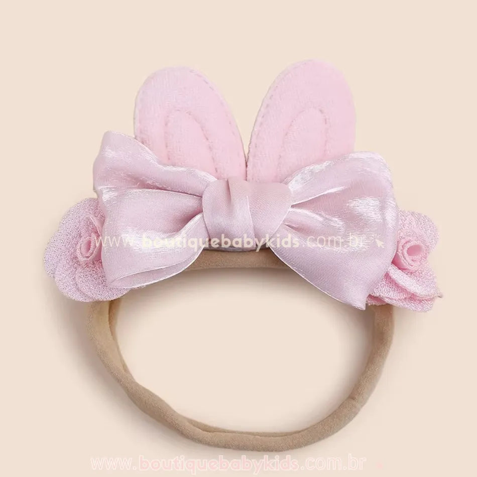 Faixa infantil rosa com orelhinhas de coelho acolchoadas e laço de cetim no centro, decorada com pequenas flores laterais.