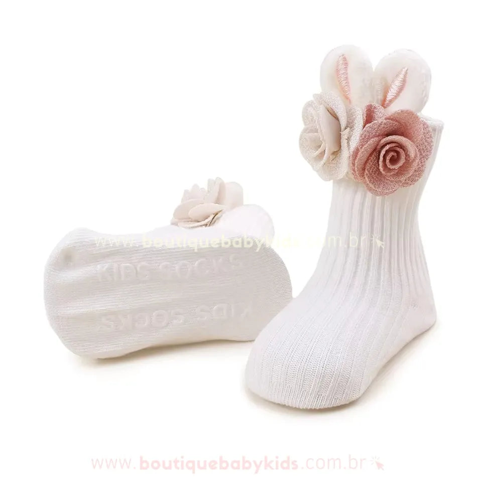 Par de meias brancas infantis em tecido canelado, com detalhe de orelhinhas de coelho e pequenas flores aplicadas na lateral, trazendo um charme especial. As solas são antiderrapantes, garantindo mais segurança para os pequenos.