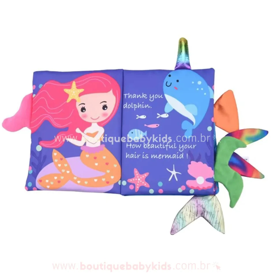 Página aberta do livro sensorial com ilustração de uma sereia de cabelos cor-de-rosa ao lado de um golfinho azul sorridente. O fundo apresenta elementos marinhos como estrelas-do-mar, peixes e corais. As caudas de tecido colorido aparecem nas laterais.