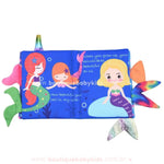 Página do livro sensorial exibindo três sereias em tons vibrantes, com caudas coloridas e detalhes subaquáticos. As frases em inglês compõem a história, incentivando a leitura e o aprendizado da língua.