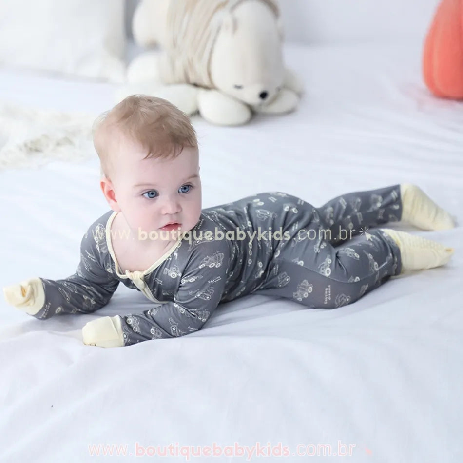 Bebê deitado com brinquedo ao fundo, usando o macacão com estampa de caminhões e tratores.