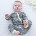 Bebê deitado segurando o pezinho, sorridente, vestindo o macacão cinza com detalhes em amarelo.