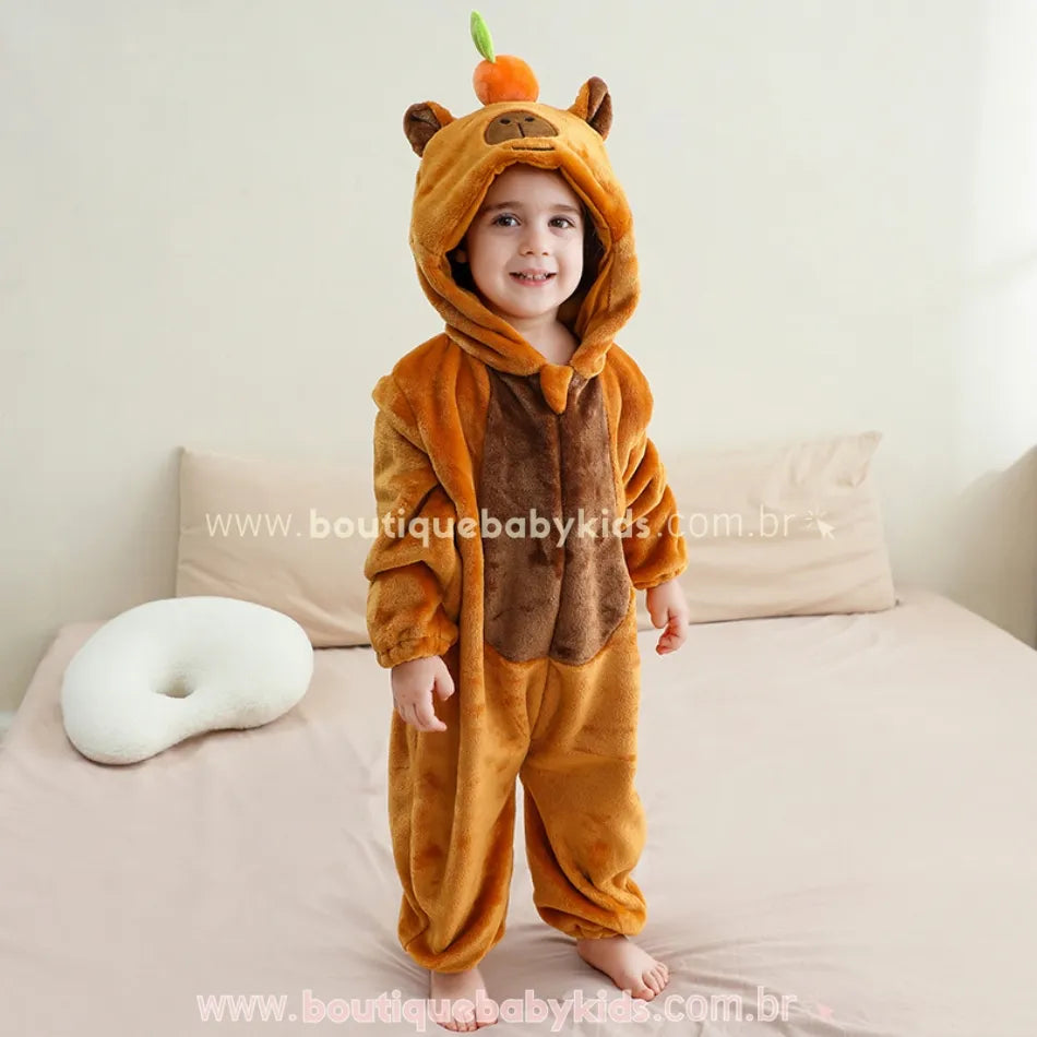 Criança em pé sobre a cama usando o Macacão Capivara do Cerrado, mostrando a parte frontal com capuz de rostinho bordado e o detalhe marrom no centro.
