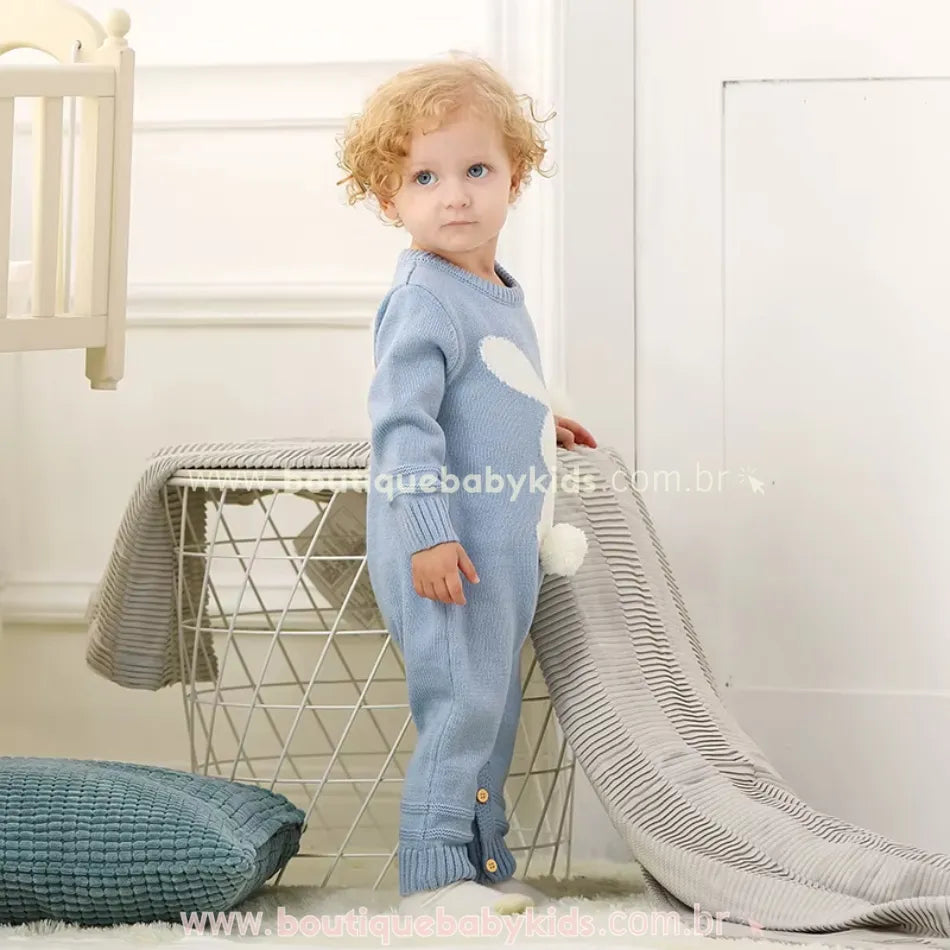 Bebê posando ao lado de um cesto, usando o Macacão Bebê Tricot Coelhinho azul. O fechamento entre as pernas por botões facilita a troca de fraldas. 