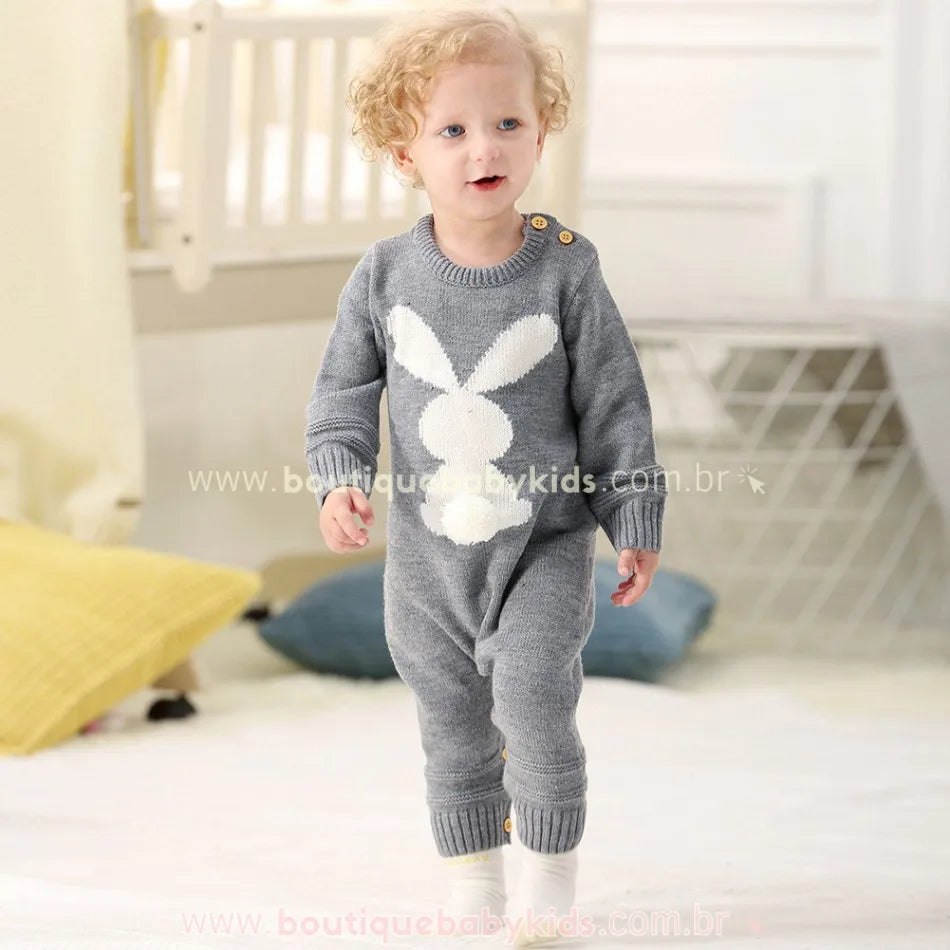Bebê em pé no quarto, vestindo o Macacão Bebê Tricot Coelhinho cinza. O design apresenta um coelhinho branco com rabinho pompom na frente, mangas longas e punhos canelados. 