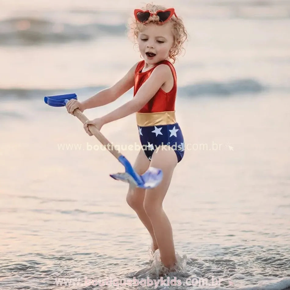 Menina loira brincando na praia com uma pá azul, vestindo o Maiô Infantil Heroína Mulher Maravilha, que possui parte superior vermelha, faixa dourada e parte inferior azul com estrelas brancas.
