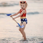 Menina loira brincando na praia com uma pá azul, vestindo o Maiô Infantil Heroína Mulher Maravilha, que possui parte superior vermelha, faixa dourada e parte inferior azul com estrelas brancas.