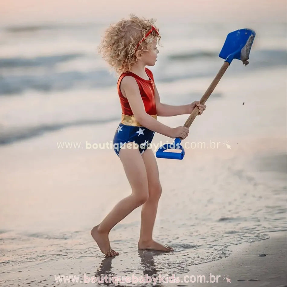Menina sorridente brincando na água do mar enquanto usa o Maiô Infantil Heroína Mulher Maravilha, segurando uma pá de brinquedo azul e vestindo um óculos vermelho.