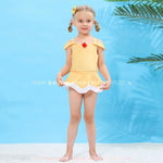 Menina loira com tranças e lacinho amarelo vestindo o maiô infantil amarelo inspirado na Princesa Bela, posando de frente com um fundo azul claro e plantas tropicais ao fundo.