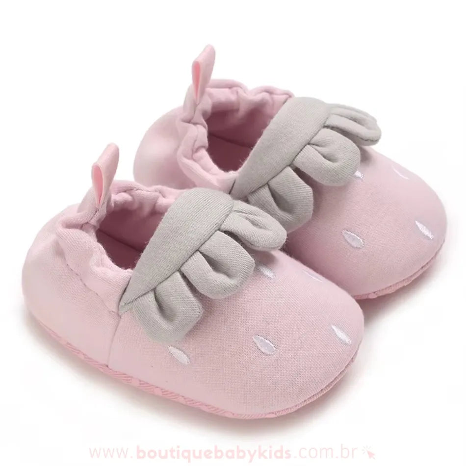 Par de pantufinhas de bebê cor de rosa com detalhe em cinza, inspirado no formato de um morango, com pequenas sementes brancas bordadas e um babado na parte superior. 