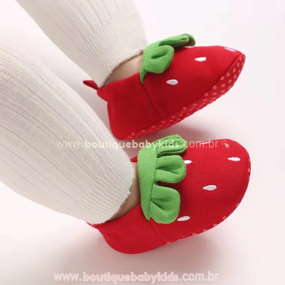 Pantufinhas temáticas de morango sendo usadas por um bebê com meias brancas, mostrando a modelagem fofa e confortável. 