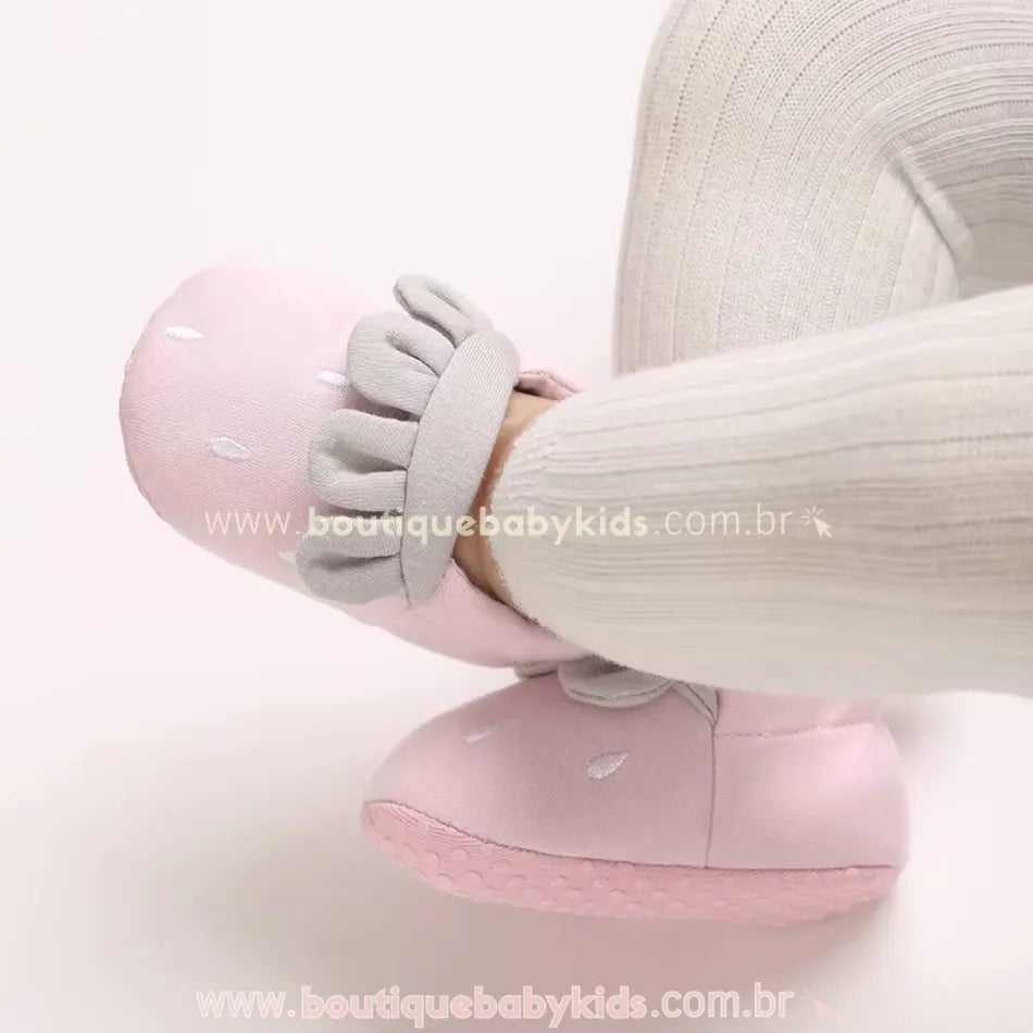 Pantufinha rosa de moranguinho no pezinho do bebê, evidenciando a sola antiderrapante e o design delicado com detalhes bordados. 