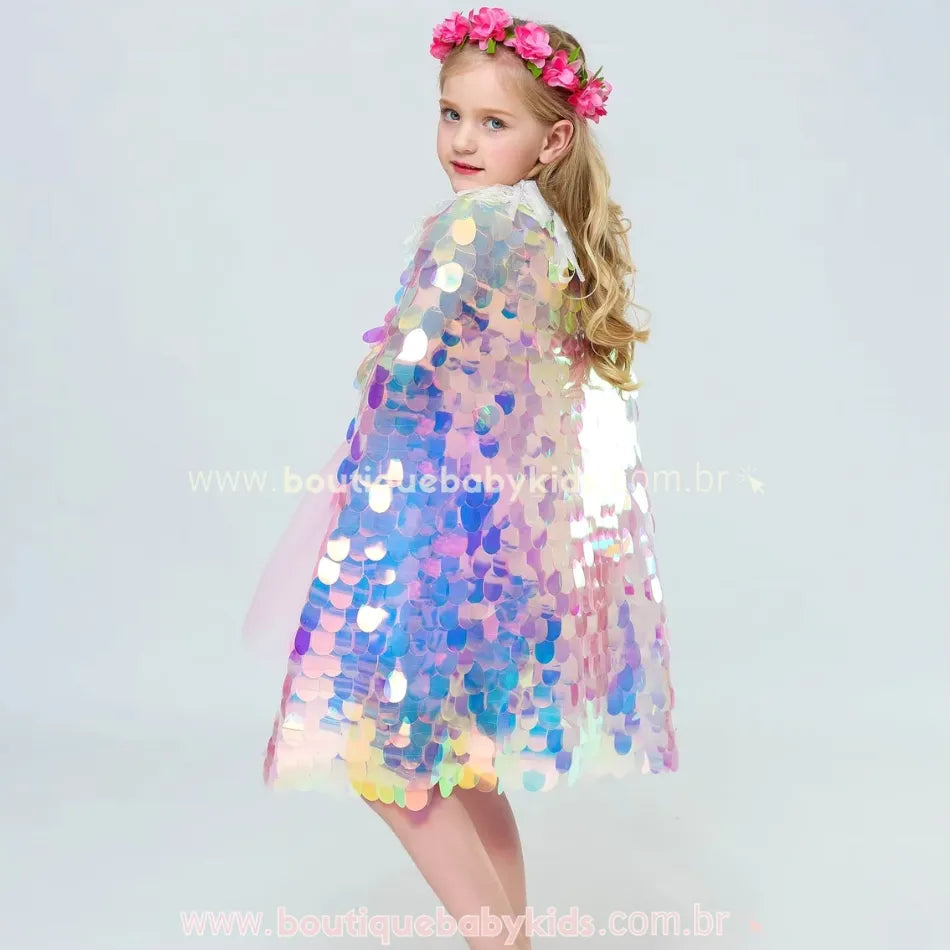 Vídeo mostrando uma criança vestindo a Capa Infantil Temática Sereia Encantada. A capa tem paetês holográficos brilhantes que refletem tons de azul, lilás e rosa. A menina gira e movimenta a capa, mostrando seu efeito cintilante. O detalhe da gola delicada com amarração dourada também aparece em destaque. O vídeo realça o caimento leve e esvoaçante da peça, perfeita para fantasias e brincadeiras temáticas.