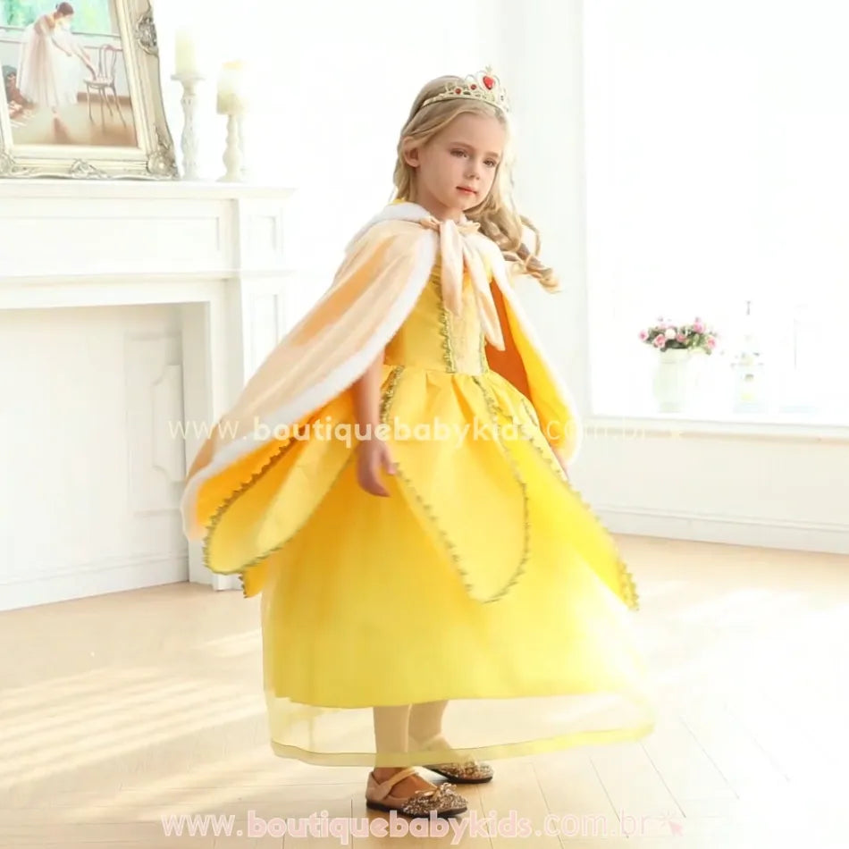 Menina vestida como uma princesa usa a capa infantil dourada com acabamento em pelúcia branca. Ela gira suavemente em um ambiente elegante, destacando o caimento fluído da peça e os detalhes sofisticados do tecido aveludado. O capuz e o laço frontal complementam o visual encantador, tornando a capa perfeita para ocasiões especiais e fantasias temáticas. 