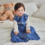 Bebê sentado na cama sorrindo, vestido com o Saco de Dormir Bebê Constelação dos Sonhos. Ao fundo, um urso de pelúcia e uma cama bem arrumada criam um ambiente aconchegante.