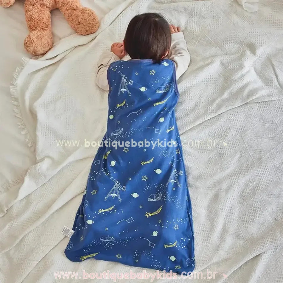 Bebê deitado de bruços na cama usando o Saco de Dormir Bebê Constelação dos Sonhos, destacando a estampa azul com elementos espaciais, como estrelas, planetas e telescópios.