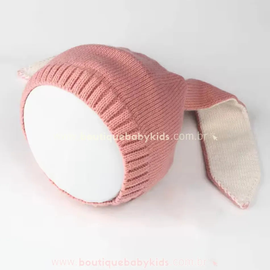 Touca de tricot infantil em tom rosé, com orelhinhas compridas de coelho e forro branco, disposta sobre uma base arredondada branca.