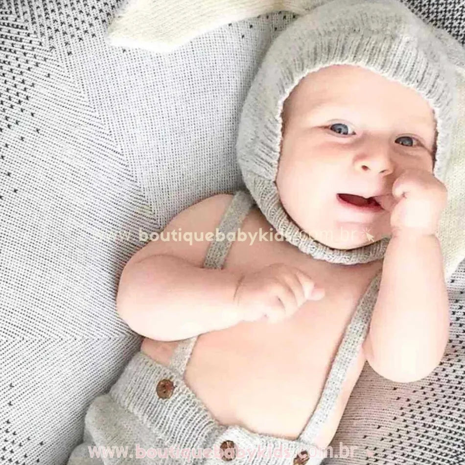 Bebê deitado sobre manta de tricot cinza, usando a touca de coelhinho e um conjuntinho suspensório combinando, com um sorrisinho no rosto.