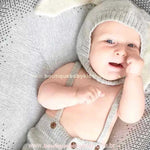 Bebê deitado sobre manta de tricot cinza, usando a touca de coelhinho e um conjuntinho suspensório combinando, com um sorrisinho no rosto.