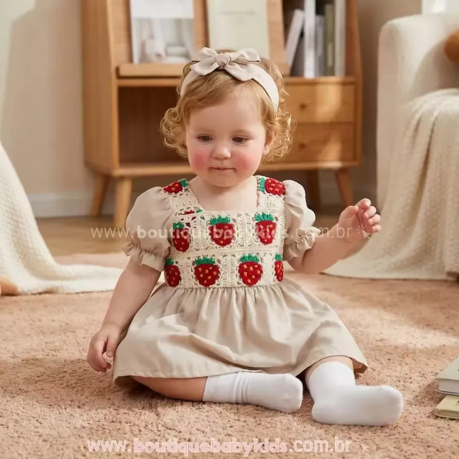 Bebê vestindo o Vestido Bebê Crochet Moranguinho com Faixa Bege sentada em um tapete bege