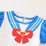 Detalhe da gola e da estampa frontal do vestido Sailor Moon, apresentando um laço vermelho com asas brancas e coração dourado, além de listras azuis que remetem ao uniforme da personagem.