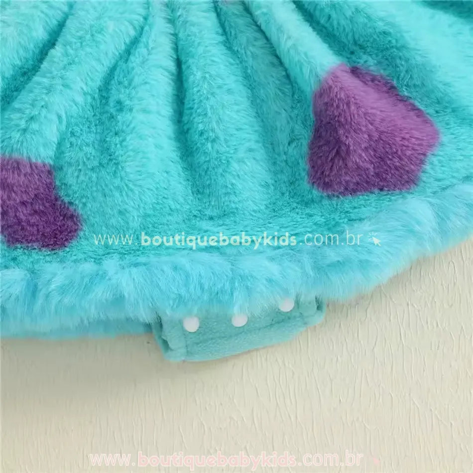 Detalhe da parte inferior do vestido bebê Sulley, com fecho de botões de pressão e tecido peluciado azul claro com manchinhas roxas.
